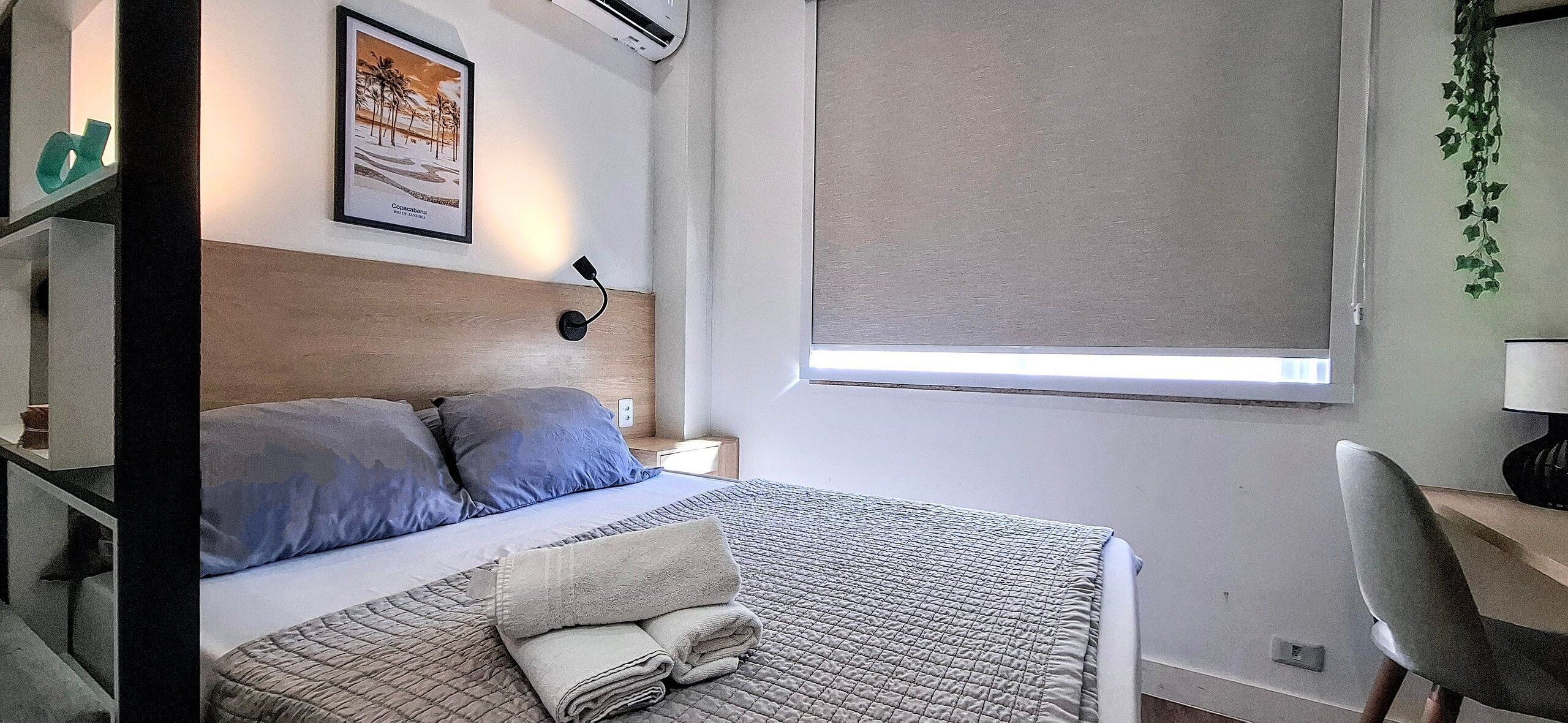A neatly made bed with a quilted bedspread and pillows, a bedside table with a lamp, a framed picture on the wall, and a window with a blind.