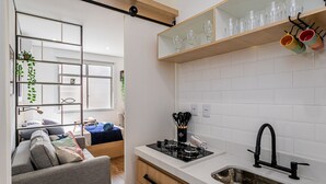 Cocina compacta con estufa a gas, un lavamanos y una encimera. Se ve un sofá y una zona de comedor al fondo.