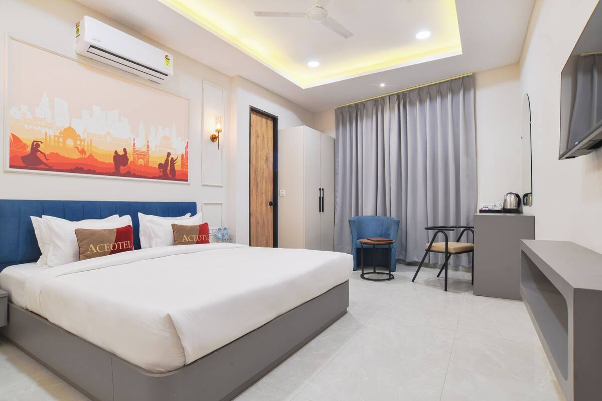 A hotel room with a bed, a desk, a chair, a flat-screen TV, an air conditioner, and a painting on the wall.