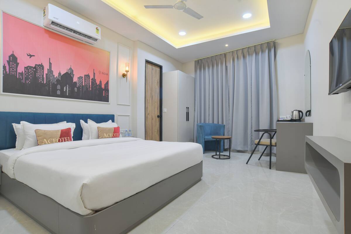 A hotel room with a bed, a desk, a chair, a television, an air conditioner, and a cityscape painting.