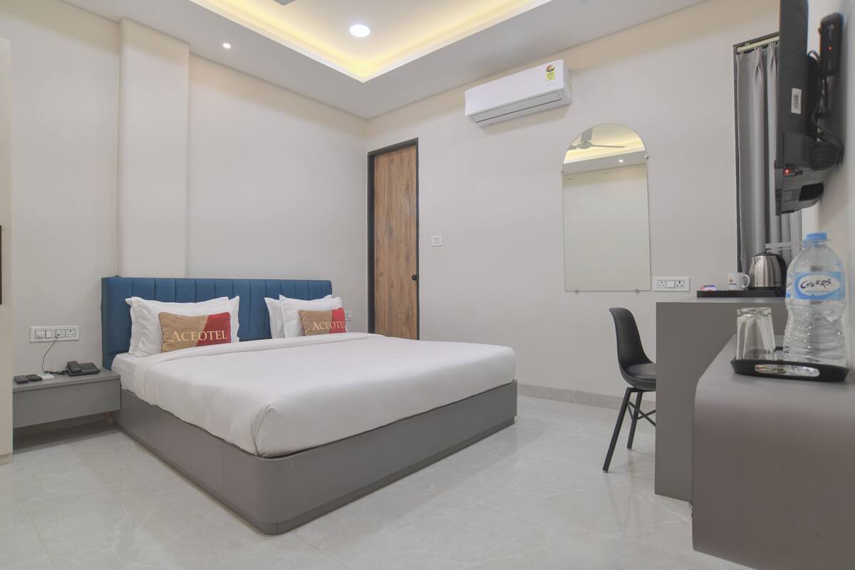 A hotel room with a bed, a desk, a chair, an air conditioning unit, and a television.