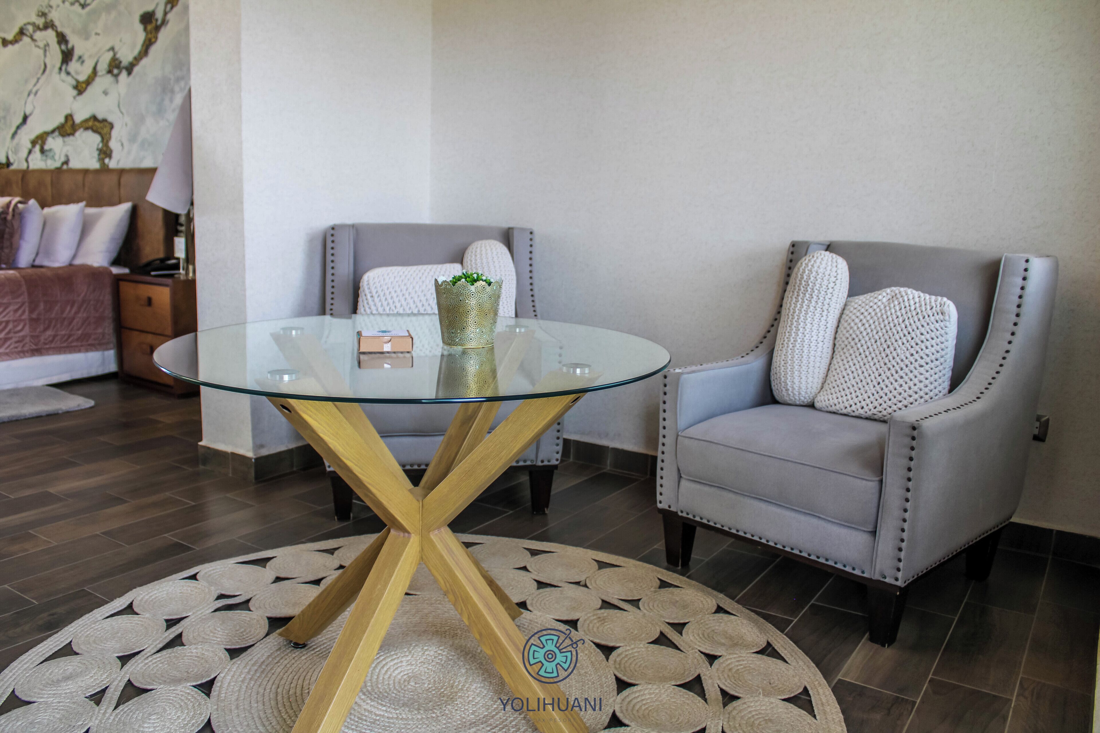 A modern living room with a glass-top coffee table, two grey armchairs, a patterned rug, and a bed with a headboard.