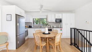 A modern kitchen with white cabinets, a stainless steel refrigerator, a microwave, and a dining area with wicker chairs and a wooden table. - La Luce - Coastal Retreat in Port Stephens (Corlette)