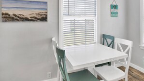 A room with a white table and chairs, a beach painting, a window with blinds, and a sign on the wall. - St-Navy Point Cottage (Pensacola)