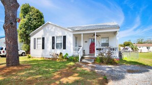 A white single-story house with a porch, a red door, and a tree in the foreground. - St-Navy Point Cottage (Pensacola)