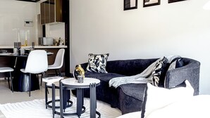 Un salón moderno con un sofá oscuro, sillas blancas, una mesita redonda y una cocina al fondo.