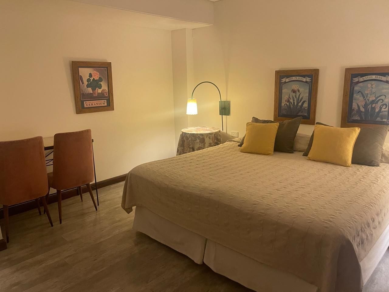A hotel room with a bed, a desk, two chairs, a lamp, and two framed pictures on the wall.