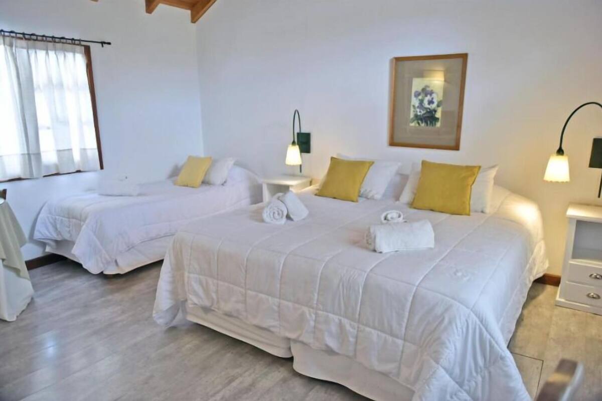 Habitación de hotel con dos camas, cada una con ropa de cama blanca y almohadas amarillas. Hay mesitas de noche con lámparas, un cuadro en la pared y una ventana con cortinas.