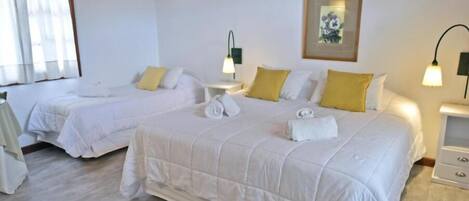 Habitación de hotel con dos camas, cada una con ropa de cama blanca y almohadas amarillas. Hay mesitas de noche con lámparas, un cuadro en la pared y una ventana con cortinas.