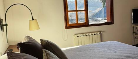 A bedroom with a bed, pillows, a lamp, a window with a view, and a radiator.
