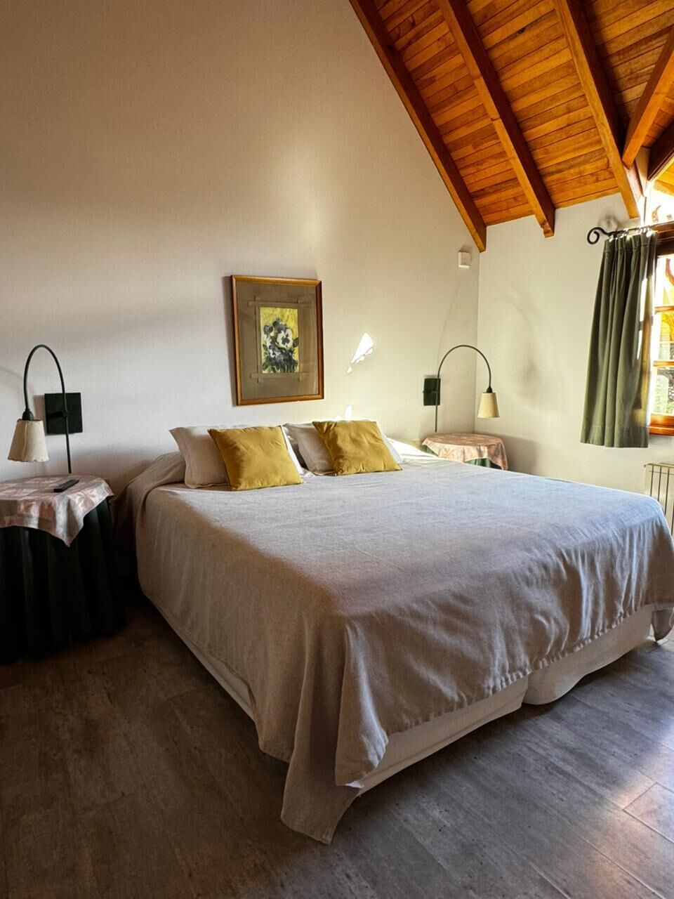 A bedroom with a large bed, two bedside tables, a nightstand, a painting on the wall, and a window with curtains.