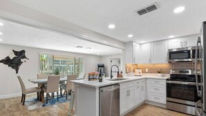 A modern kitchen with white cabinets, stainless steel appliances, and a dining area with a table and chairs. - 4BR Waterfront | Boat Dock + Pool | 11-Min Walk to Beach | Sleeps 8 (Hudson)