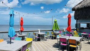 A deck with outdoor seating, a view of the ocean, and string lights. - 4BR Waterfront | Boat Dock + Pool | 11-Min Walk to Beach | Sleeps 8 (Hudson)