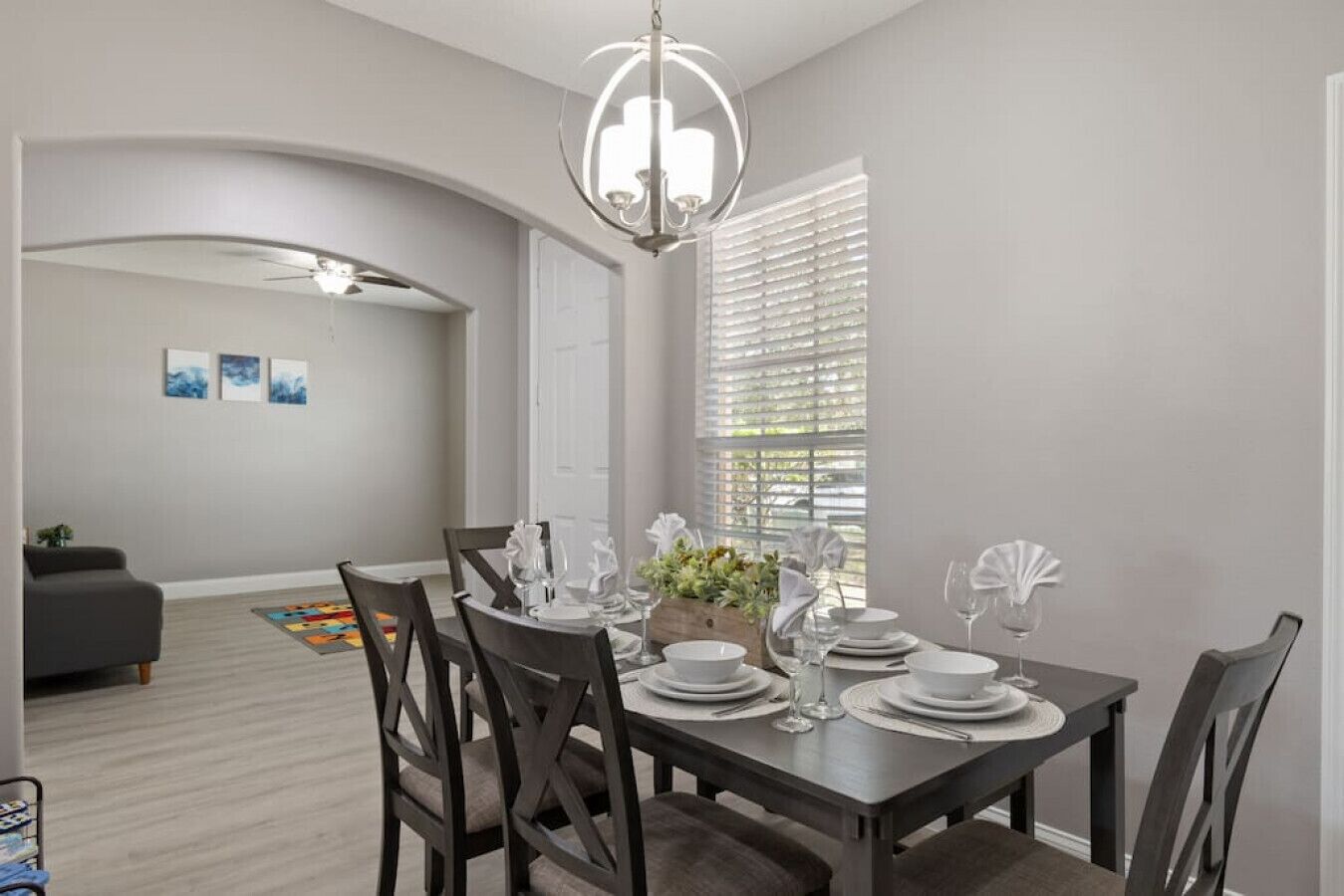 A dining area with a dark wooden table set for four, white plates, and glassware. There is a chandelier above the table and a window with blinds to the right.