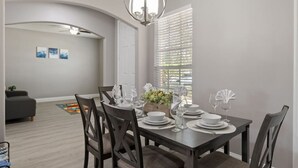 Un comedor con una mesa de madera oscura dispuesta para cuatro personas, platos blancos y cristalería. Sobre la mesa, un candelabro y, a la derecha, un ventanal con persianas.