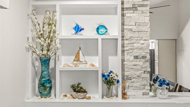 Ein weißes Regal mit Dekoration, darunter eine blaue Vase, ein Modell eines Segelboots, Muscheln und eine kleine Pflanze.