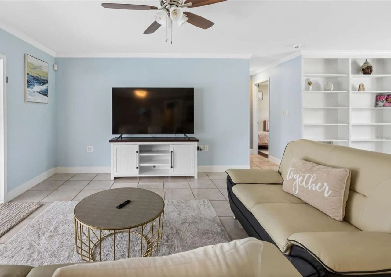 Un salón con un televisor de pantalla plana, un sofá beige con una almohada que dice 'juntos', una mesa de centro redonda y un mueble de entretenimiento blanco.