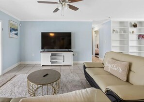 A living room with a flat-screen TV, a beige sofa with a 'together' pillow, a round coffee table, and a white entertainment unit. - 3BR | Saltwater Pool + Spa | Solar Heated | 9 Min to Airport | Sleeps 7 (Clearwater)