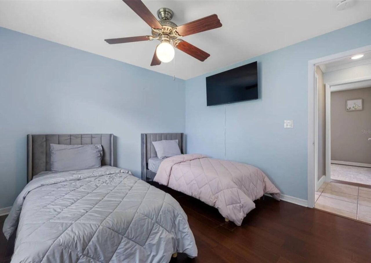 Une chambre avec deux lits, un ventilateur de plafond et un téléviseur à écran plat fixé au mur.