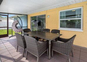 A covered patio with a table and chairs, a hanging chair, and a view of a house through glass doors. - 3BR | Saltwater Pool + Spa | Solar Heated | 9 Min to Airport | Sleeps 7 (Clearwater)