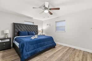 Un dormitorio con cabecera acolchada, cama con colcha azul, mesita de noche con lámpara y ventilador de techo.