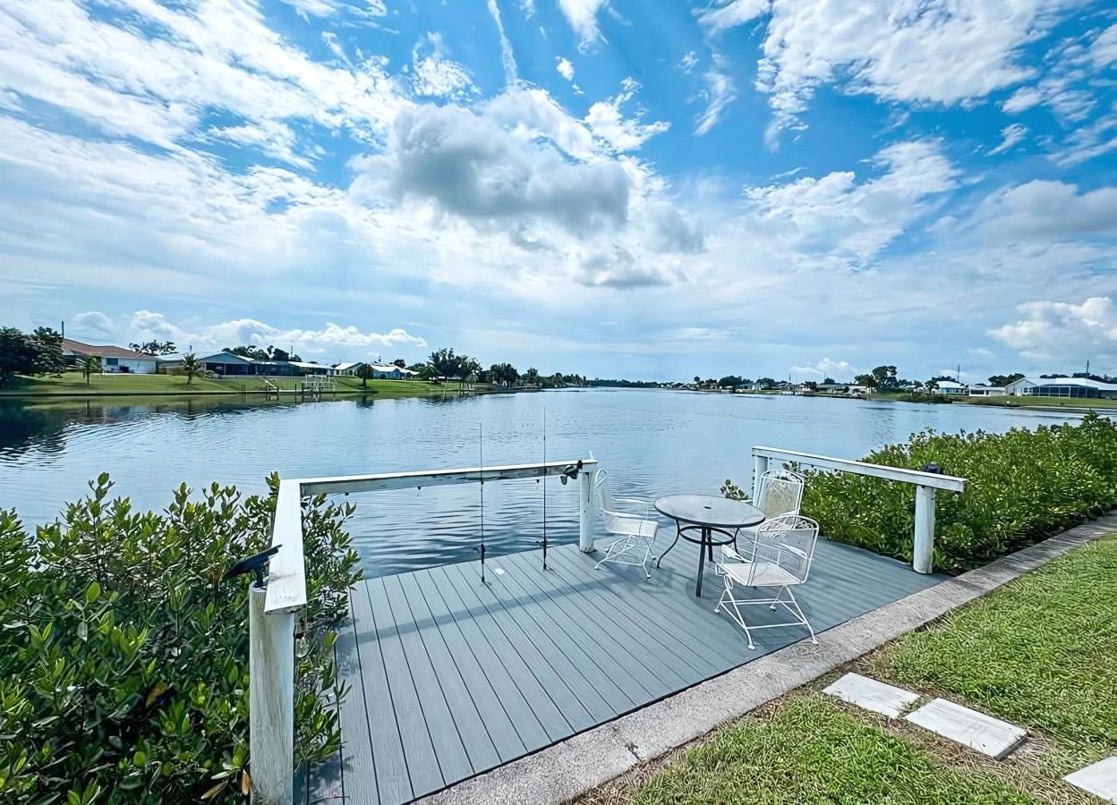Una terraza de madera con una mesa y sillas da vista a un lago tranquilo, rodeado de follaje y un cielo despejado.
