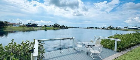 Una terraza de madera con una mesa y sillas da vista a un lago tranquilo, rodeado de follaje y un cielo despejado.