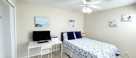 A bedroom with a bed, a desk, a chair, a ceiling fan, and artwork on the walls.