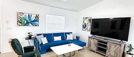 Ein modernes Wohnzimmer mit einem blauen Sofa, einem weißen Couchtisch, einem Holz-TV-Schrank und einem Deckenventilator.