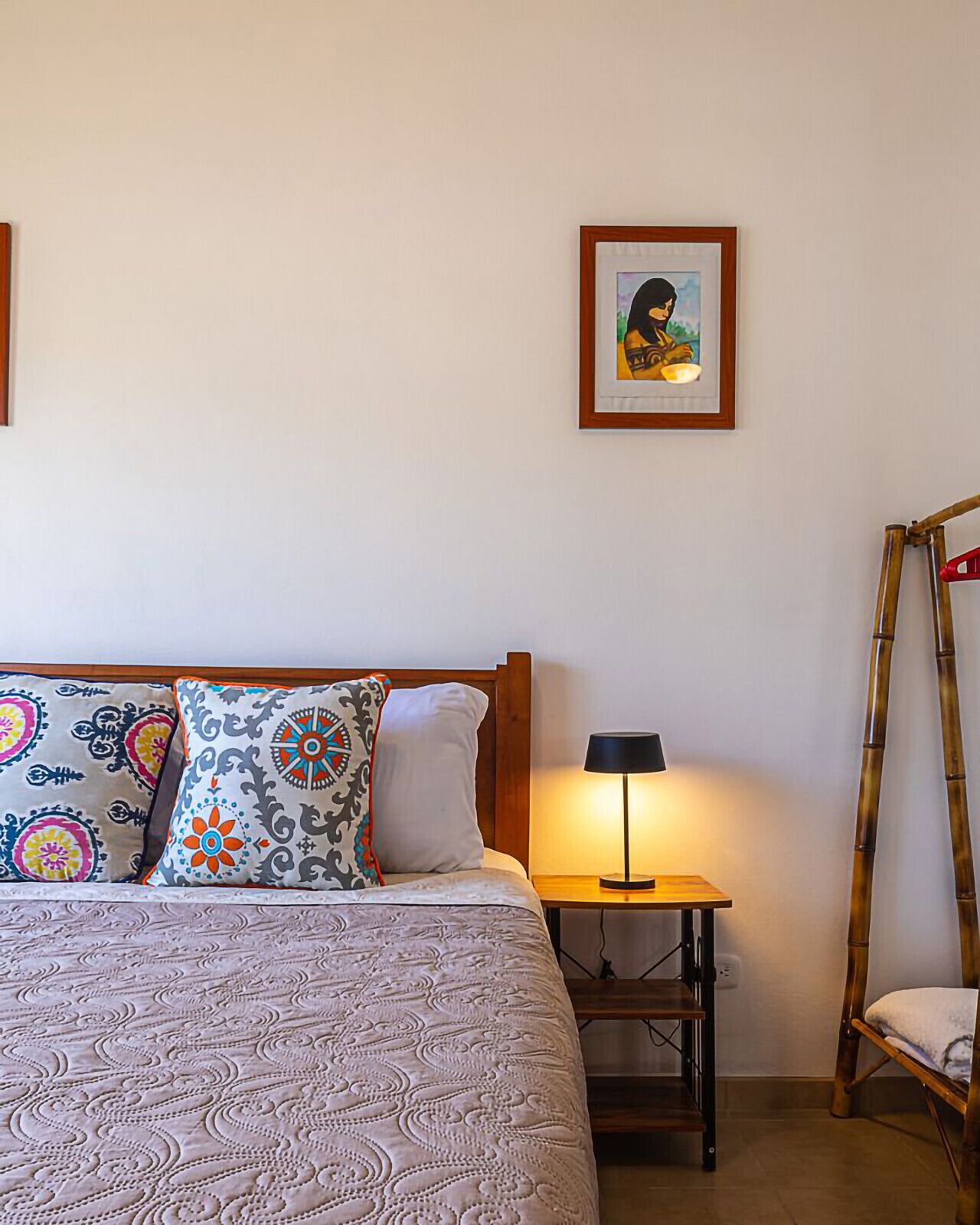 A neatly made bed with patterned bedding, a bedside table with a lamp, a framed picture on the wall, and a wooden ladder against the wall.