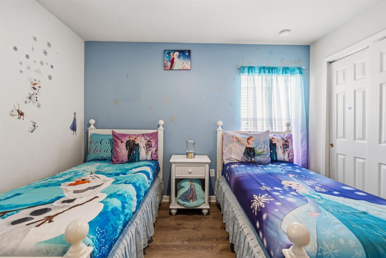 A bedroom with two beds, each with a Disney-themed bedspread, a nightstand, a small table, and a window with curtains.