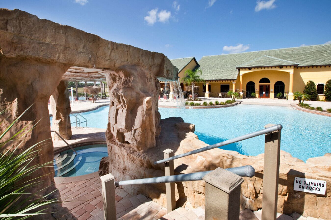 A pool area with a natural rock formation, a no-climbing-on-rocks sign, and a building with a tiled roof in the background.