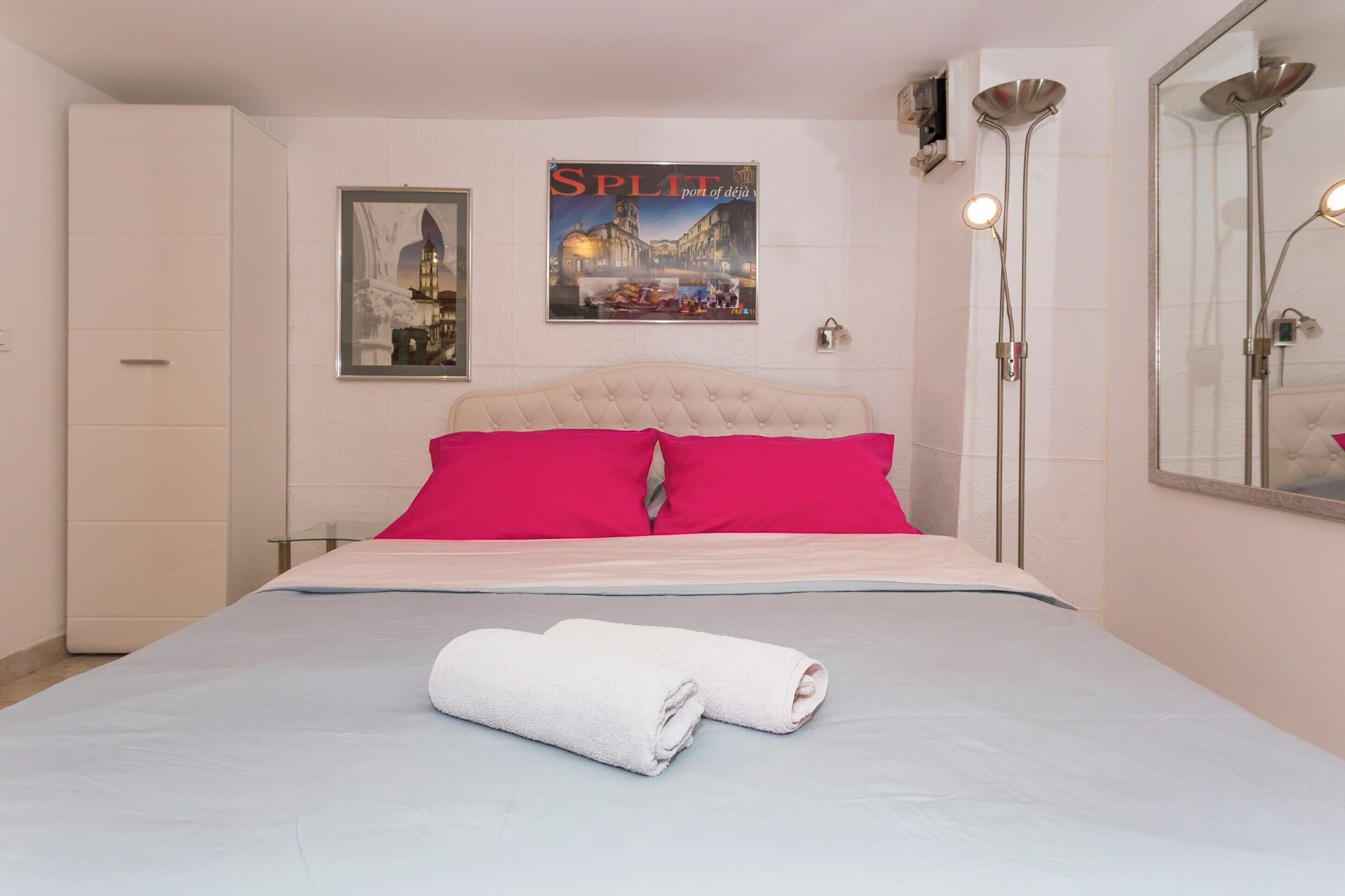 Una cama bien hecha con edredón blanco, dos almohadas rosa brillante y una toalla blanca enrollada sobre una colcha azul claro.