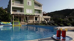 Una piscina con sillas de descanso, un edificio verde con balcones y una montaña al fondo.