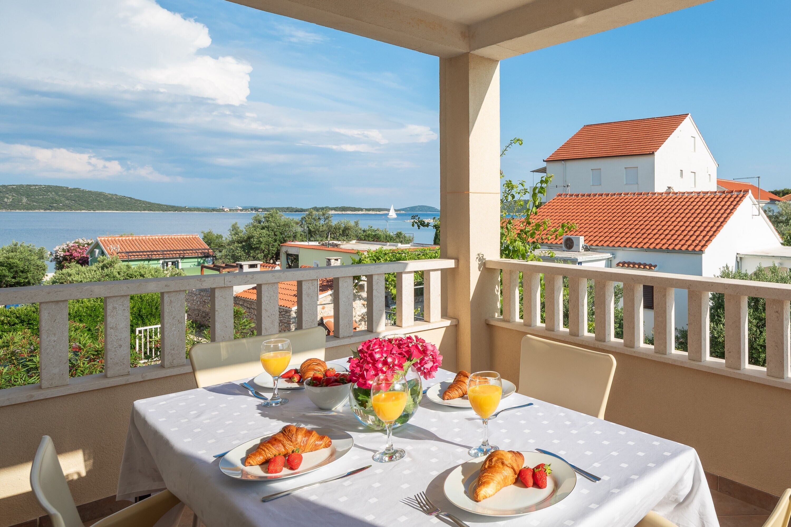 A balcony with a table set for breakfast, overlooking a scenic view of a lake and buildings.