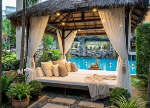 Ein Pavillon mit Strohdach, einem Bett, einem Pool und tropischen Pflanzen.