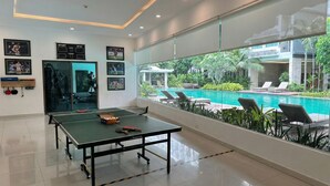 A ping pong table with paddles and balls, a wall with framed sports photos, and a view of a swimming pool and greenery. - Laguna beach resort maldives 3 - Modern studio pool view with balcony in Jomtien (Muang Pattaya)