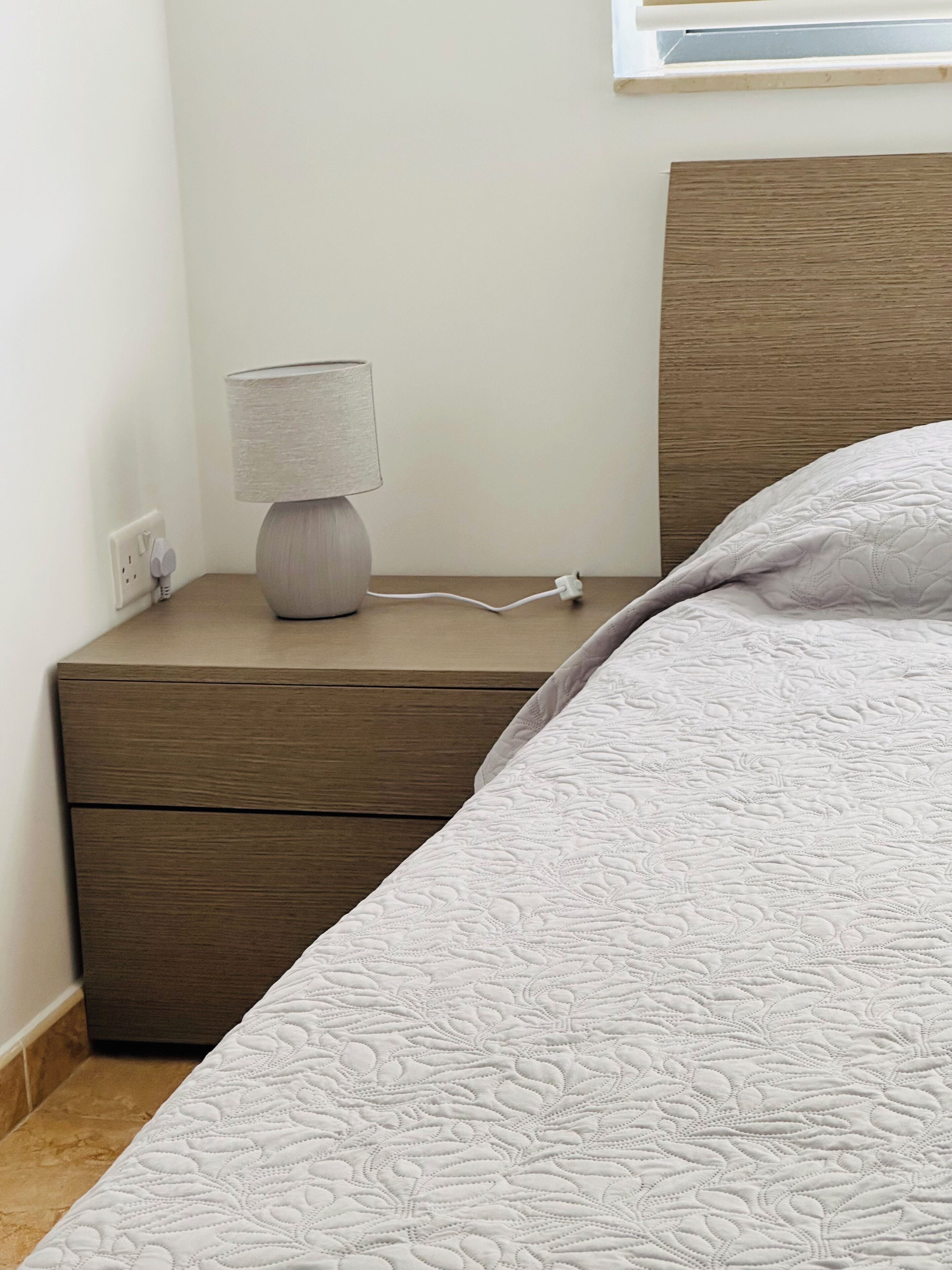 Un lit bien fait, recouvert d’une housse blanche matelassée, une lampe de chevet avec un abat-jour blanc et une table de chevet en bois dotée d’un tiroir.