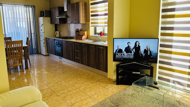 Una cocina moderna con armarios de madera oscura, estufa integrada y microondas. Hay una zona de comedor con mesa y sillas, y un televisor que muestra 'Law & Order' en la pared.