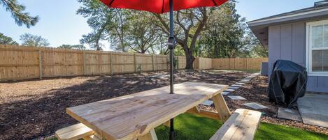 Una mesa de picnic de madera con bancos, un paraguas rojo y una barbacoa en un patio trasero.