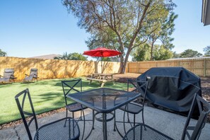 Un patio trasero con mesa de picnic, sillas, una parrilla y una sombrilla.