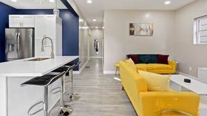 A modern kitchen with white cabinets and stainless steel appliances, a bar area with stools, and a bright yellow sofa in a living area with a painting on the wall. - Chic Design and Modern Comfort - Inviting Boho Escape (Indianapolis)