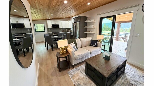 Een moderne keuken met witte kasten, een eethoek met tafel en stoelen, een bank en een bijzettafeltje. Er hangt een grote spiegel aan de muur en er is uitzicht op het buitenlandschap door een schuifdeur van glas.