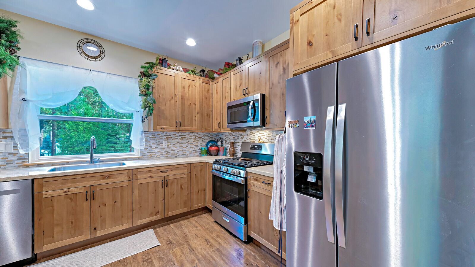 A modern kitchen with wooden cabinets, stainless steel appliances, a window with a view of greenery, and a microwave above the stove.