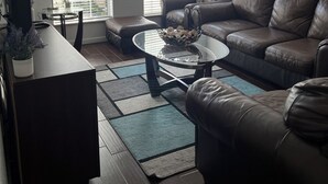 A living room with a brown leather couch, a glass coffee table, and a TV on a stand. There are two windows with blinds, a ceiling fan, and a painting on the wall. - 3 Bedroom 3 Bath Townhome Cul-de-Sac (Tallahassee)