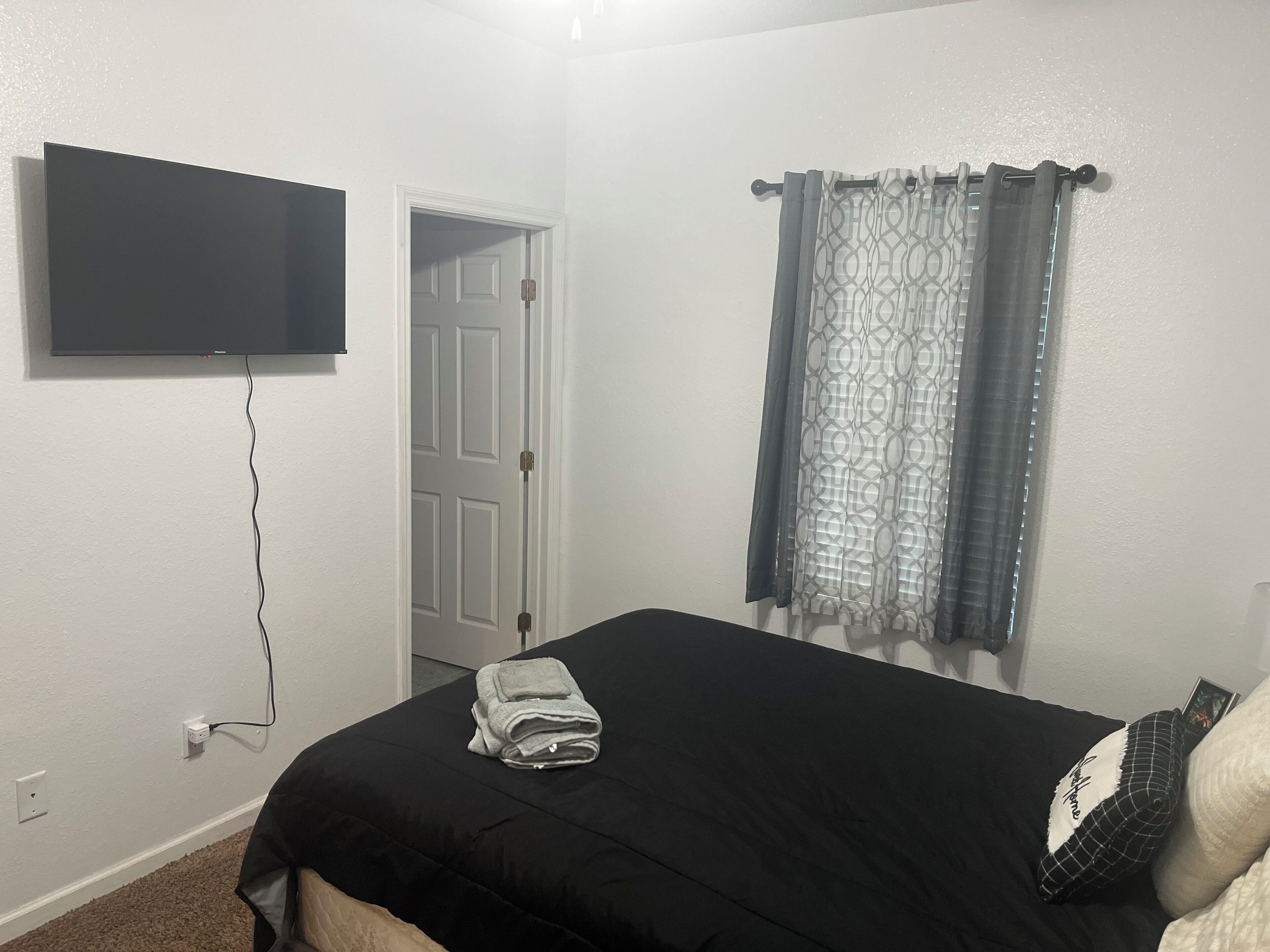 Un dormitorio con cama, televisor de pantalla plana en la pared, una puerta y una ventana con cortinas estampadas.