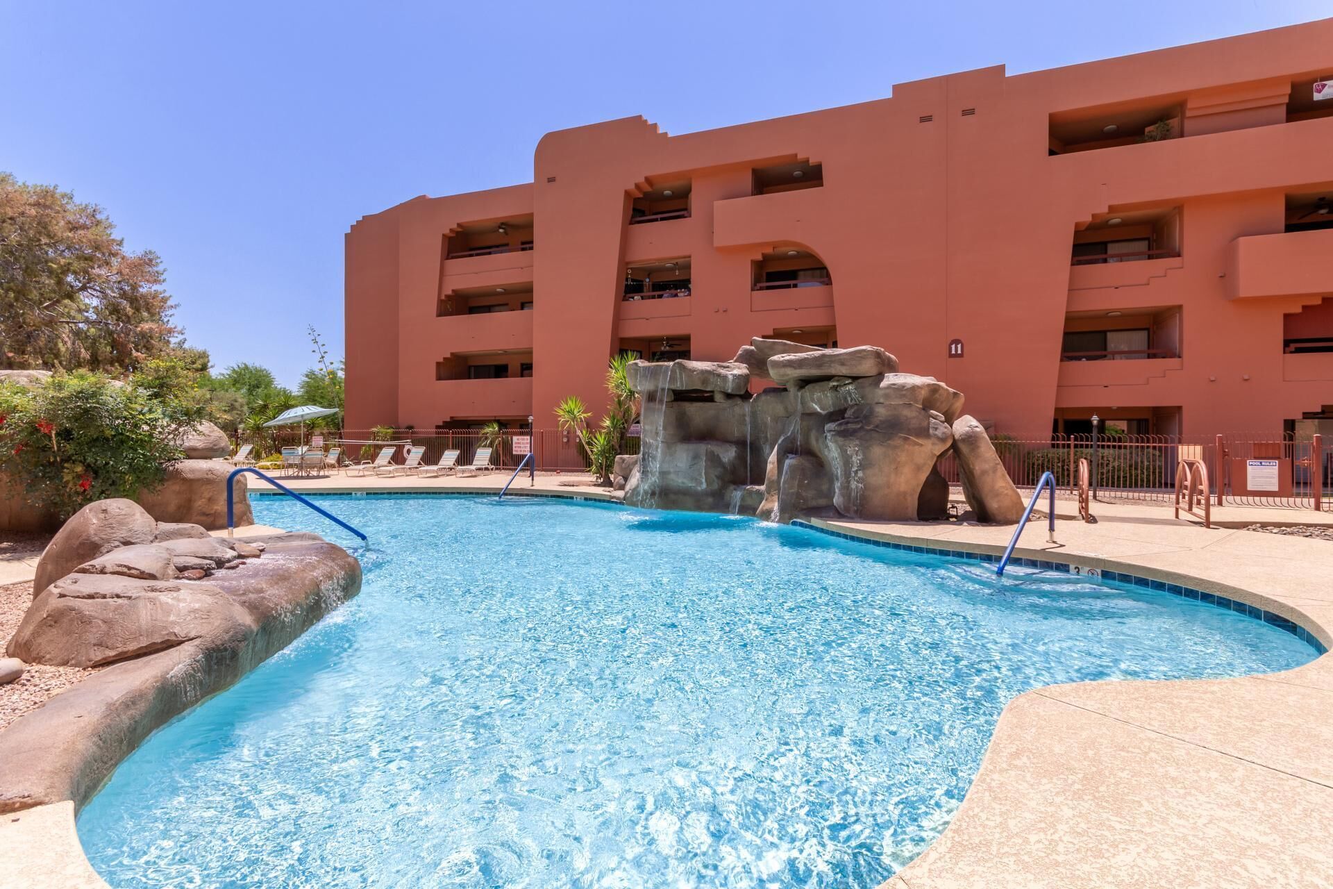 A swimming pool with a rock feature and lounge chairs, surrounded by a building with multiple balconies.