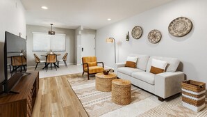 A modern living room with a white sofa, a dining table with chairs, a TV, and decorative woven baskets.