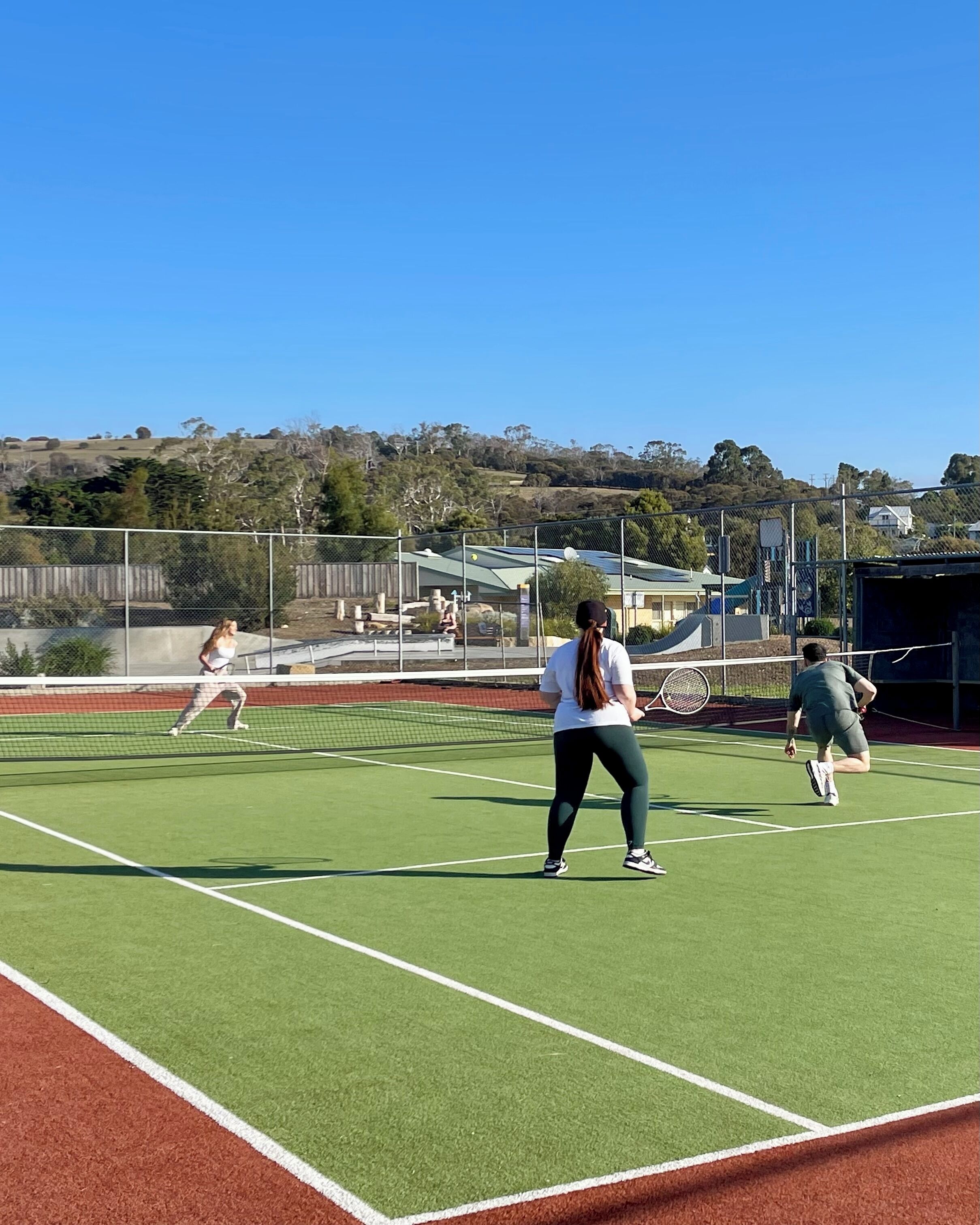 A tennis court with players, a fence, and a building in the background.