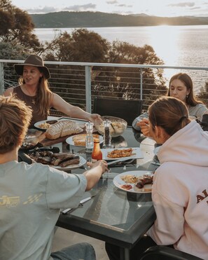 Un grupo de personas cenando al aire libre, con una mesa preparada para la comida, una botella de salsa y una vista de agua al fondo.